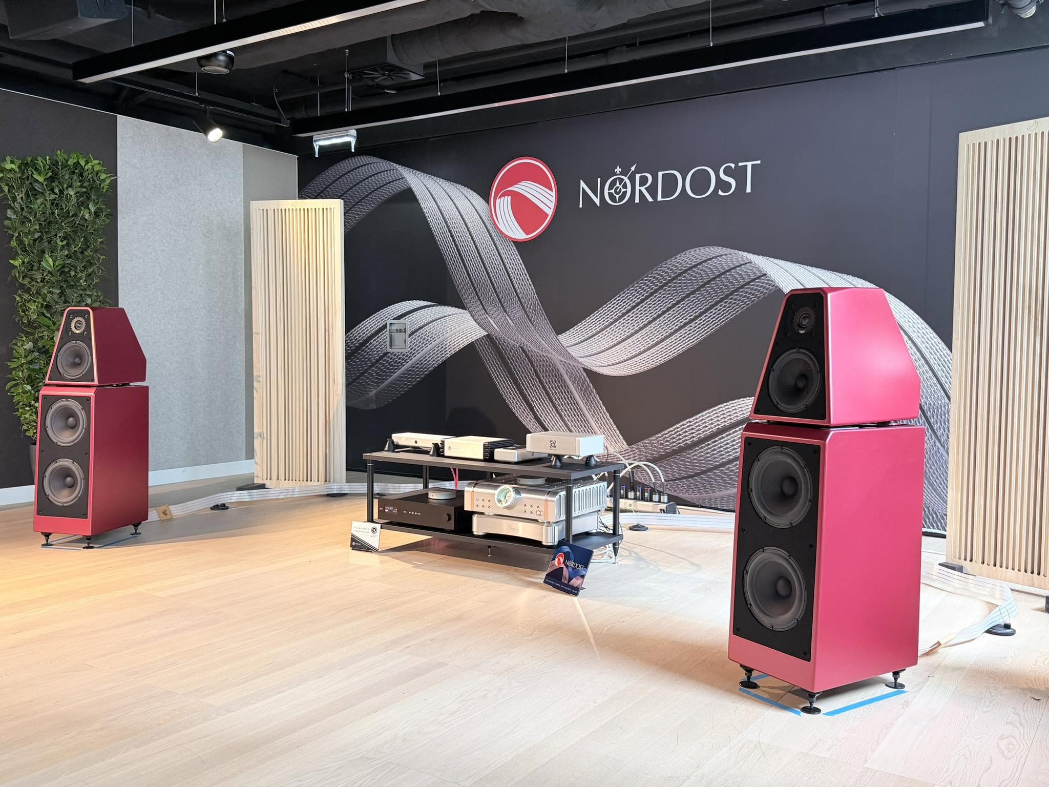 Showroom with pink floor-standing speakers, audio gear on a rack, and a Nordost banner on a dark wall in the backstory.