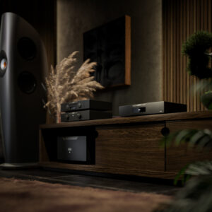 Dimly lit home audio room with a tall black speaker on the left, wooden wall panels, and AV equipment on a shelf right of center.